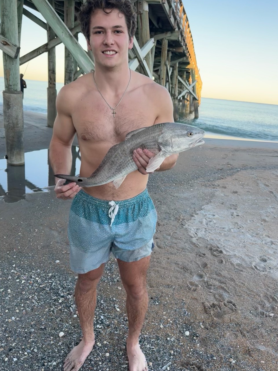 Ian holding a fish on the beach beneath a pier